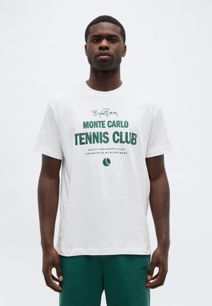 Νεαρός άνδρας φοράει ένα λευκό T-shirt "Monte Carlo Tennis Club" και πράσινα σορτς, στέκεται μπροστά σε έναν απλό λευκό φόντο.