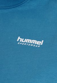 Nærbillede af blå stof med hvidt "hummel SPORTSWEAR" logo trykt på brystområdet af en beklædningsgenstand.