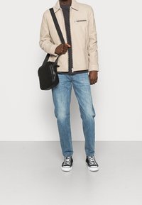 Beige Lederjacke mit Reißverschlusstaschen, schwarzes T-Shirt, hellblaue Jeans und schwarze Sneakers mit weißen Sohlen sowie eine schwarze Umhängetasche.
