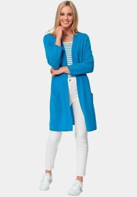 Langer blauer Cardigan aus weichem Stoff, mit geradem Schnitt, zwei vorderen Taschen und langen Ärmeln. Kombiniert mit weißen Hosen und Sneakers.