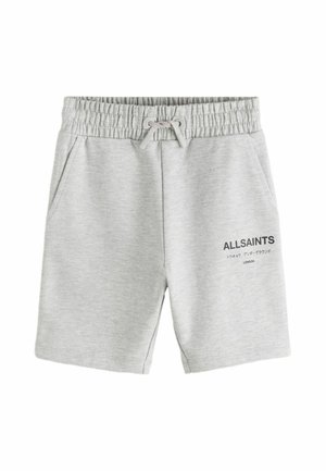 Teenager iført en lysegrå hættetrøje og matchende shorts med hvide sokker og hvide sneakers, der står foran en ensfarvet grå baggrund.
