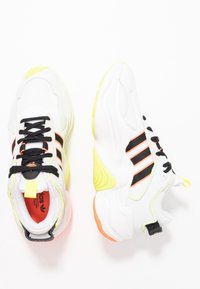 Vita Adidas-sneakers med svarta ränder, svarta snören, gula och orange accenter, visade från ovan och sidan på en vit bakgrund.