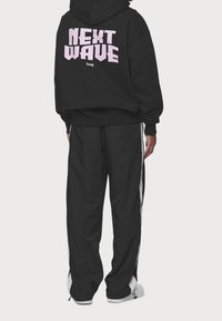 Persona che indossa un hoodie nero con la scritta "NEXT WAVE" in rosa sulla schiena e pantaloni sportivi neri con strisce bianche sui lati, in piedi su uno sfondo bianco.