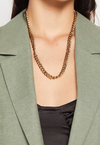 Zlatý řetězový náhrdelník s propletenými články, nošený přes černý oděv, doplněný světle zeleným blejzrem. Hladký kovový povrch.