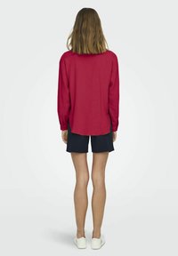 Red long-sleeve shirt made of textured fabric, paired with navy shorts. Features a curved hem and relaxed fit. Neutral background.