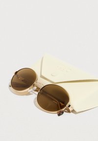 Barna, kerek napszemüveg aranyszínű fémszerkezettel és áttetsző díszítésekkel, egy világos sárga, zárható fedelű védőtokon pihen.