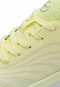 Gros plan d'une chaussure de sport jaune pâle avec une tige en mesh texturé, des lacets croisés et des motifs ondulés subtils sur le côté.