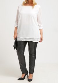 Top blanco de manga larga con escote cuadrado, combinado con pantalones de cuero negros ajustados y tacones negros de punta, sosteniendo un bolso de mano negro.