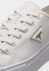 Baskets en toile blanche avec des lacets blancs et un logo triangulaire en métal Guess USA sur le côté près de l'orteil.