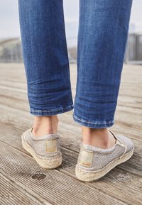 Zilveren espadrilles met een bovenkant van geweven stof en jute zool. Platte ontwerp met ronde neus en een klein logo-accent op de hiel. Spijkerbroek zichtbaar.