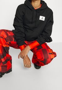 Personne accroupie portant un sweat à capuche noir avec un logo blanc et un pantalon à motifs rouges, montrant des tatouages sur les mains et des chaussures noires sur un sol blanc.