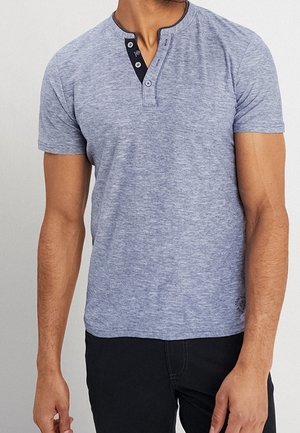 Mann trägt ein hellgraues Henley-Shirt mit kurzen Ärmeln und schwarzer Knopfleiste sowie schwarze Hose, steht vor einem einfarbigen Hintergrund.