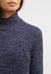 Pull à col roulé bleu marine en tissu tricoté texturé. Présente un motif côtelé et des détails subtils sur les épaules.