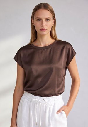 Frau mit glatten blonden Haaren, die ein kurzärmeliges braunes Satintop und weiße Hose mit Bündchen trägt, steht vor einem einfachen hellen Hintergrund.