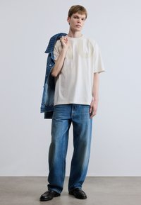 Αμάνικο εκρού μπλουζάκι μεγάλης διάστασης με μεγάλο ανοικτόχρωμο σχέδιο, σε συνδυασμό με φαρδιά μπλε τζιν παντελόνια και μπλε τζιν μπουφάν περασμένο πάνω στον ώμο.