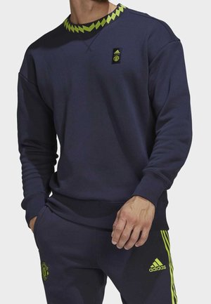 Mężczyzna w granatowej bluzie Adidas Manchester United i dopasowanych spodniach z neonowozielonymi detalami i logo, ręce swobodnie ułożone wzdłuż ciała.
