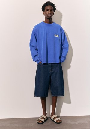 Mężczyzna stojący przy białej ścianie, ubrany w niebieską koszulę z długim rękawem, ciemne dżinsowe spodenki do kolan oraz białe sandały z dwoma paskami.