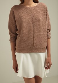 Pulover marrón texturizado con mangas largas y cuello redondo, combinado con una falda blanca de corte acampanado. Tejido suave con un ajuste holgado.