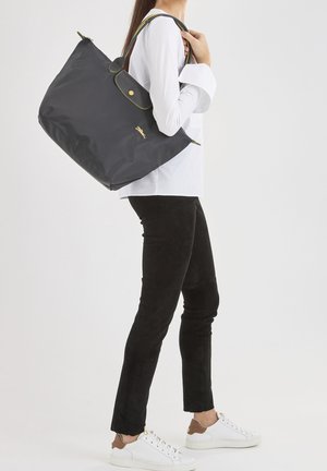 Zwarte nylon tote bag met gele accenten en een gouden logo, gedragen op de schouder. Model draagt zwarte broek en witte sneakers.