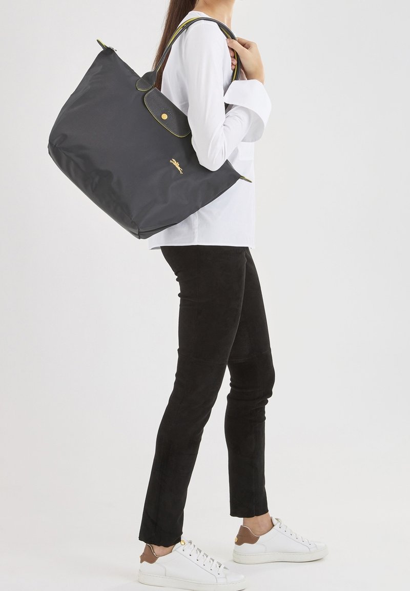 Sac fourre-tout noir en nylon avec des accents jaunes et un logo doré, porté sur l'épaule. Le mannequin porte un pantalon noir et des baskets blanches.