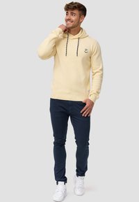 Sudadera amarilla con capucha, cordones negros y logo, combinada con pantalones ajustados azul marino y zapatillas blancas. Textura de punto plano.