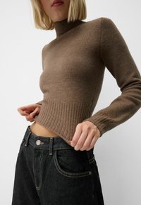 Maglione marrone a costine con collo alto, corpetto aderente e maniche lunghe, abbinato a jeans scuri a vita alta con cuciture visibili.
