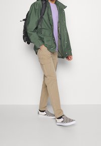 Groene lichtgewicht jas, paarse T-shirt, beige cargobroek en zwart-witte geruite instapschoenen. Zwarte rugzak met riemen.