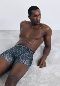 En mann uten skjorte som ligger tilbakelent på et lyst teppe iført mønstrede boxer-truser med "Björn Borg" midjebånd, ser mot høyre.