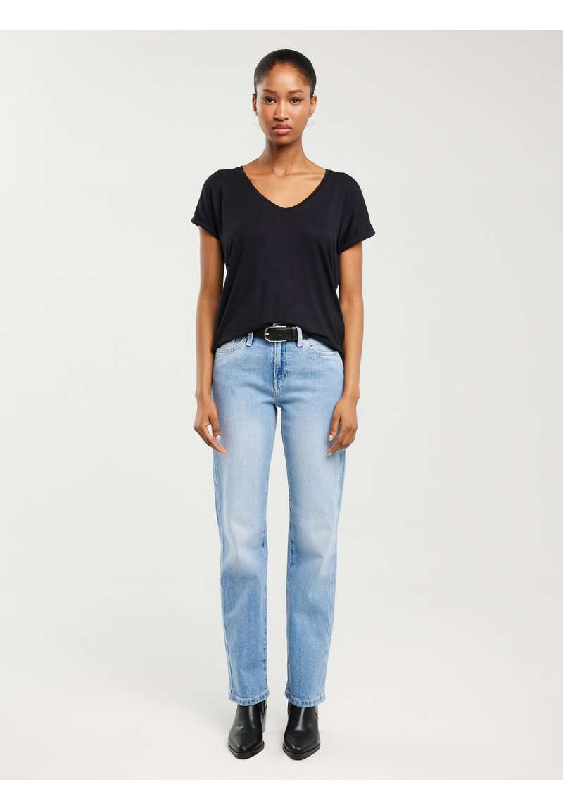 Naine seisab otse vaadata, seljas must V-kaelusega T-särk, helesinised teksad musta vööga ja mustad pahkluu saapad valgel taustal.