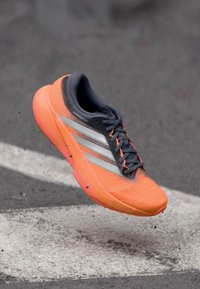 Leuchtend orangefarbener Laufschuh mit grauem Mesh-Obermaterial, drei silbernen Streifen, perforierter Textur und einer gedämpften Sohle. Leichtes Design.