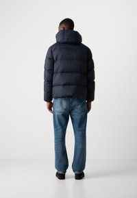 Navy puffer jacket with a high collar and horizontal quilting. Worn with light blue denim jeans and brown shoes. Back view.