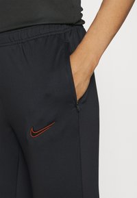 Granatowe dresy z elastycznym paskiem w talii, boczną kieszenią i pomarańczowym logo Nike. Materiał wydaje się gładki o lekko elastycznej fakturze.