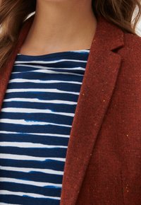 Blazer texturé de couleur rouille porté sur une chemise bleu marine à rayures blanches. Le blazer présente des taches de couleur disséminées sur tout le tissu.