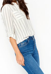 Vrouw in een witte lange mouwen shirt met zwarte strepen en opgerolde mouwen, gecombineerd met hooglopende blauwe jeans tegen een witte achtergrond.