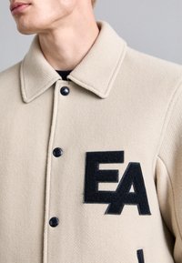 Giacca in lana beige con finitura testurizzata, bottoni neri e un grande patch ricamato "EA" nero sul lato sinistro del petto.
