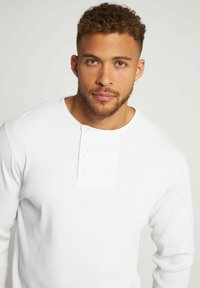 Hvid langærmet skjorte med ribbet tekstur, der har en rund halsudskæring og en lille knaplukning på brystet.