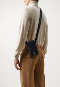 Borsa a spalla rettangolare nera con chiusura a zip, tracolla e logo verde. Indossata su pantaloni beige, su uno sfondo neutro.