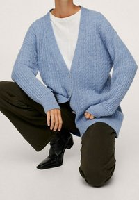 Cardigan tricoté bleu clair avec un col en V, une texture côtelée et une fermeture à boutons, porté par-dessus un haut blanc et associé à un pantalon vert foncé.