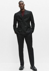 Zwart dubbelgeknoopt colbert met inkeping revers, gecombineerd met zwarte broek en zwarte loafers; gedragen over een donker bordeauxrood t-shirt.