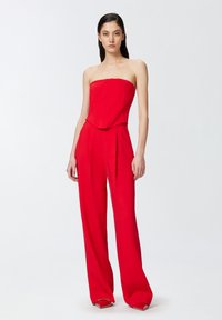 Mono rojo sin tirantes con corsé ajustado, pantalones de pierna ancha y detalles de pliegues, confeccionado en una tela suave. Presenta una silueta limpia y moderna.