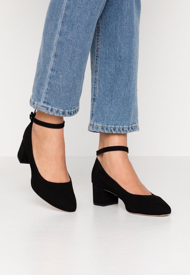 Femme portant un jean bleu clair et des chaussures à talons blocs en suède noir avec bride à la cheville, debout sur un sol blanc.