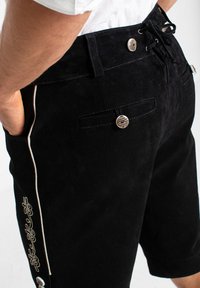 Pantaloni corti in suede neri con una texture liscia, caratterizzati da ricami laterali e una chiusura a bottone in vita. Inclusi tasche frontali e cordoncino.