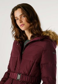 Chaqueta de plumas burdeos con capucha forrada de piel, cintura ceñida con cinturón, cierre de cremallera y textura acolchada, llevada sobre un suéter gris.