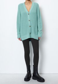 A turquoise knitted cardigan with a V-neck, long sleeves, and six large buttons, paired with black leggings and chunky black boots.