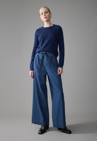 Un pull en maille bleu marine associé à un pantalon en denim à jambes larges avec une ceinture à la taille. Le mannequin porte des bottines noires à talons. Fond gris uni.