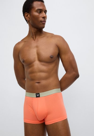 Cuecas boxer laranja com cintura verde e patch de logotipo preto. Tecido suave e design ajustado, exibidos num modelo com porte atlético.