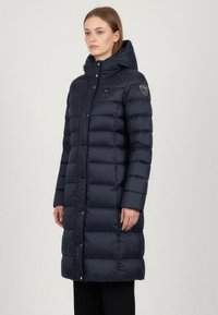 Cappotto puffer blu marino con cappuccio, caratterizzato da un design trapuntato, bottoni a scatto e una patch del logo sulla spalla sinistra. Maniche lunghe e lunghezza fino al ginocchio.