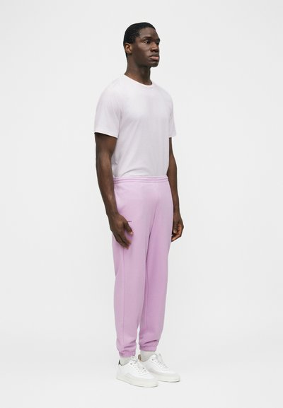 Mężczyzna stojący, ubrany w białą koszulkę z krótkim rękawem, jasnofioletowe spodnie dresowe, białe trampki, na jednolitym białym tle.