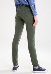 Vaqueros ajustados de color verde oliva con una textura suave, costuras de contraste y bolsillos traseros. Combinados con una camisa azul estampada y zapatillas azules.