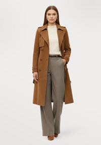 Kvinna som bär en kamelfärgad trenchcoat över en vit tröja, grå vida byxor, brunt bälte och bruna skor, stående mot en vit bakgrund.